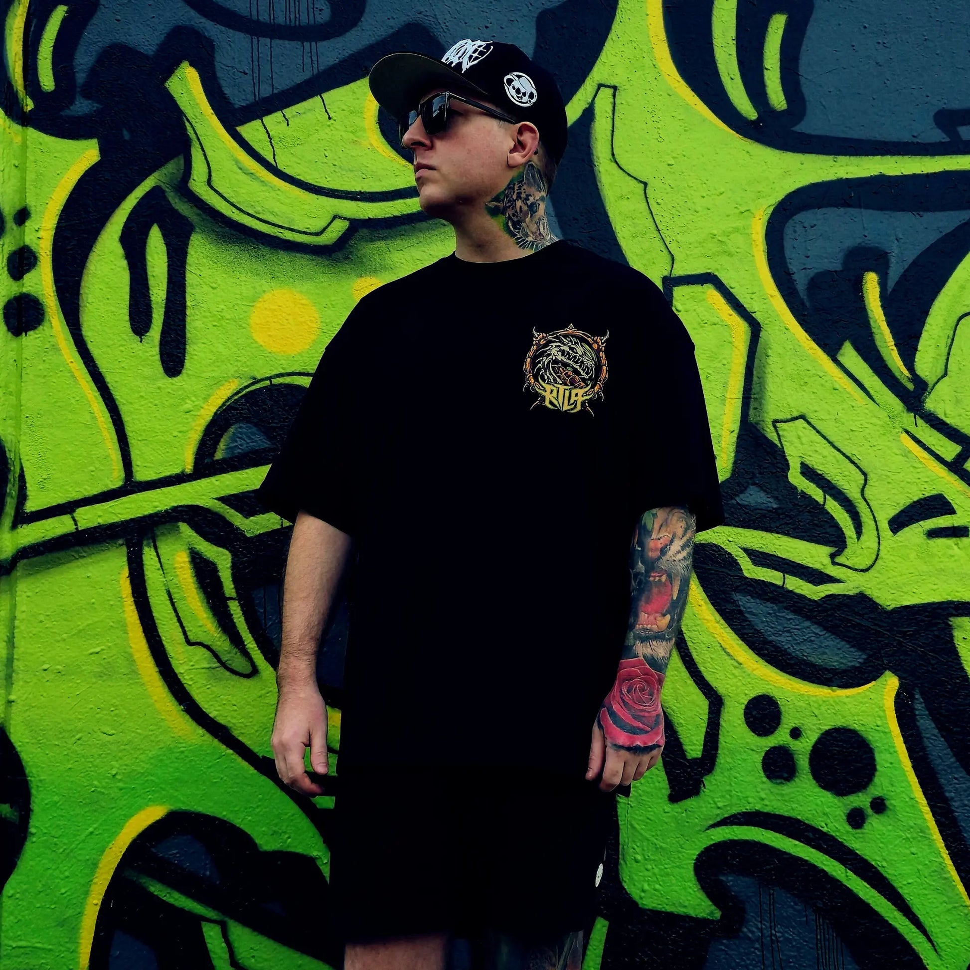 Person wearing a black t-shirt with a logo in front of a vibrant graffiti wall. RTLA-Apparel