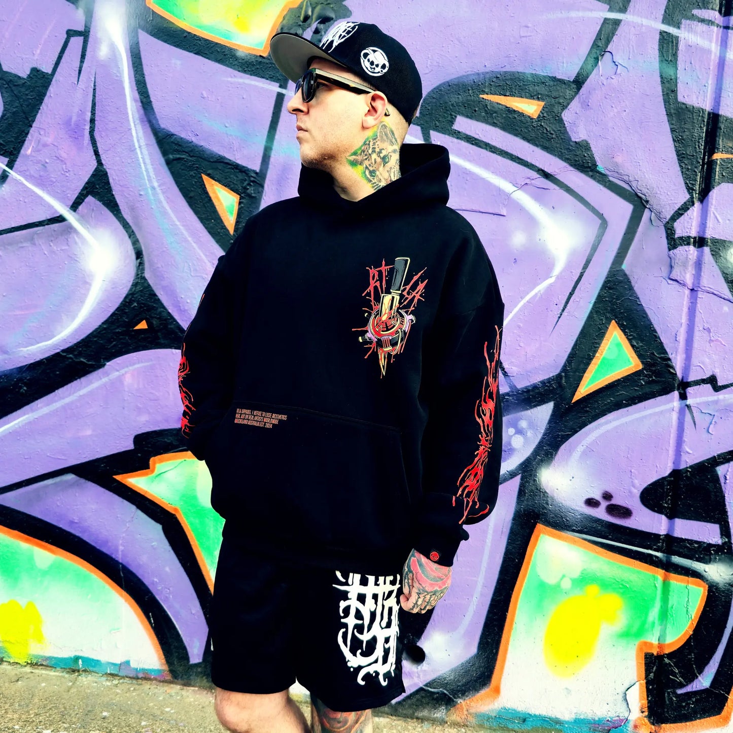 Person wearing a black hoodie with a graphic design in front of a colorful graffiti wall.