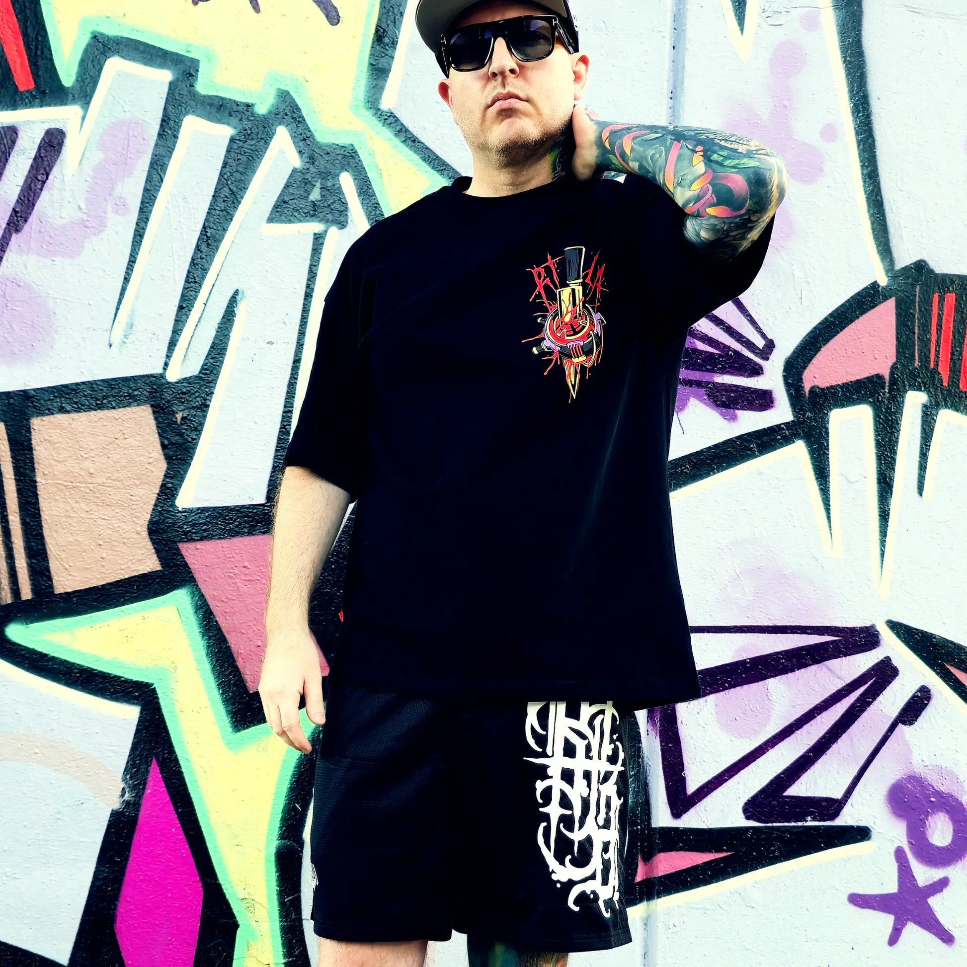 Person wearing a black t-shirt with a colorful graphic design in front of a graffiti wall.