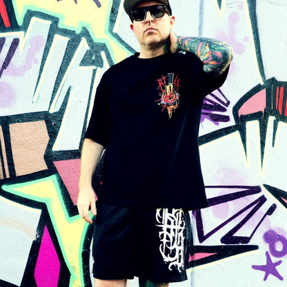 Person wearing a black t-shirt with a colorful graphic design in front of a graffiti wall.