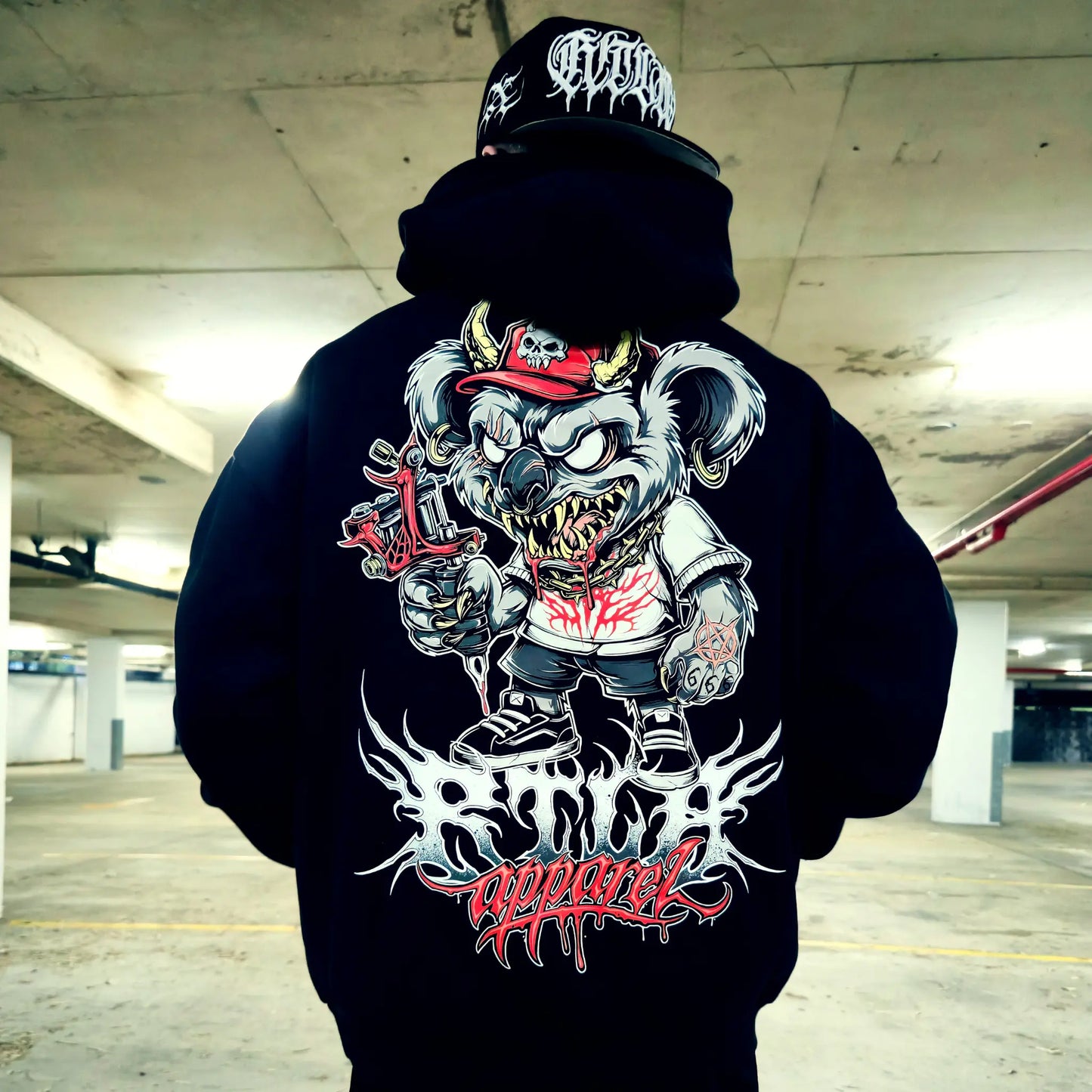 Person wearing a black hoodie with a graphic design of a bear in a red cap and white shirt, with 'Apparel' text, in an indoor setting.