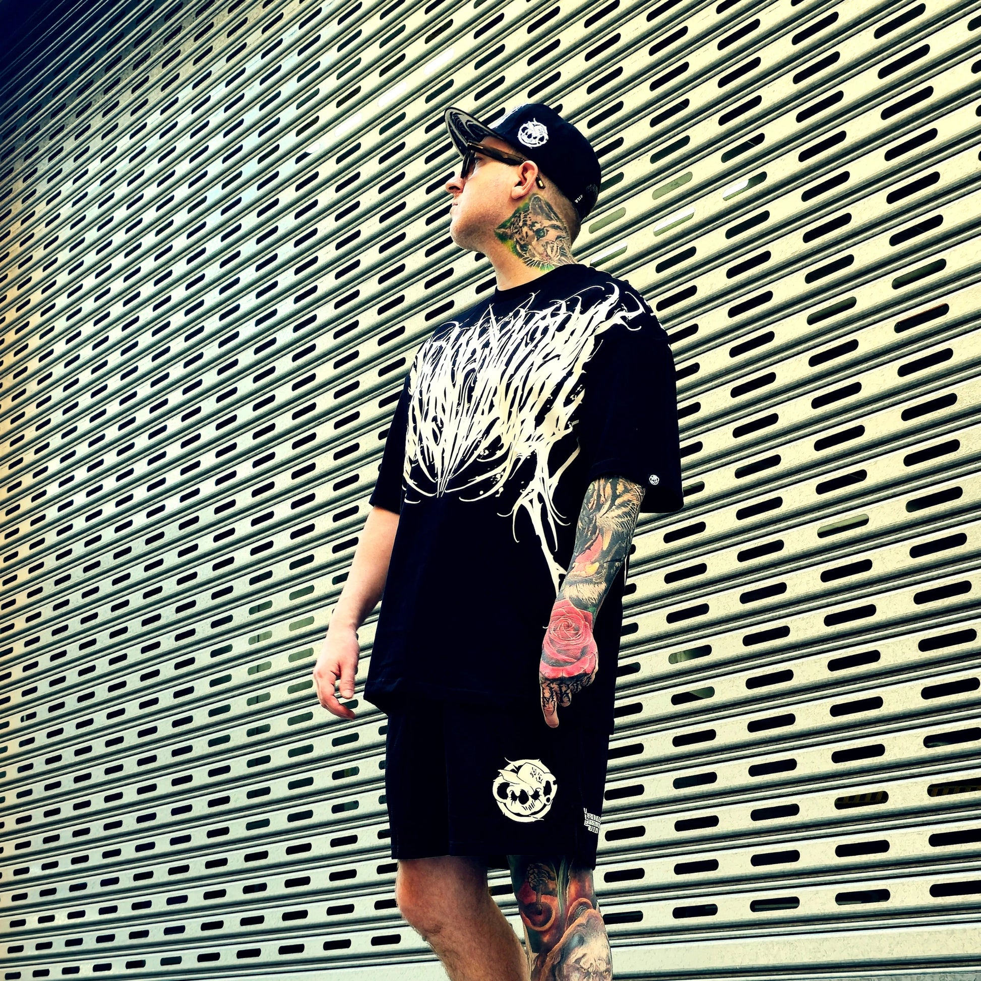 Person wearing a black t-shirt with white graphic design and black shorts, standing against a metallic shutter background.
