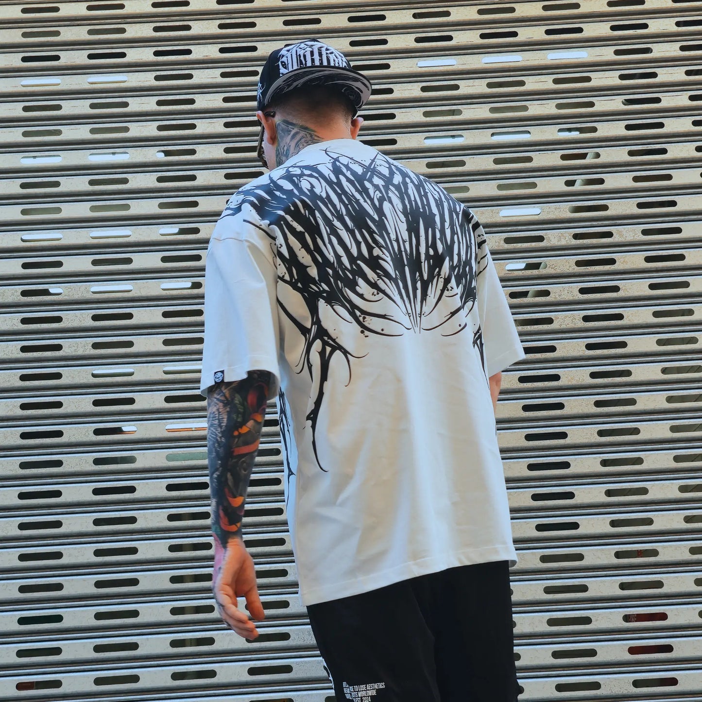 Person wearing a white t-shirt with black graphics, standing in front of a metallic shutter.