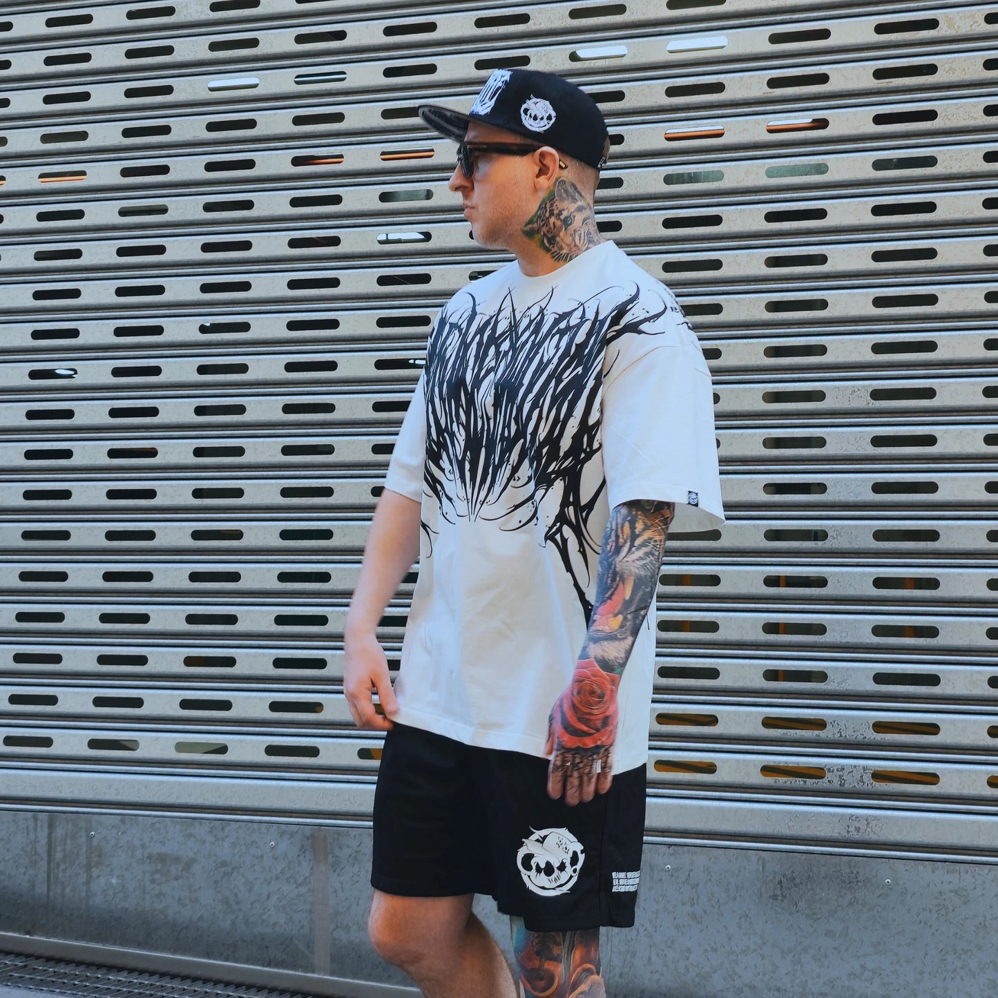 Man wearing a white t-shirt with black graphics and black shorts, standing in front of a metallic shutter.