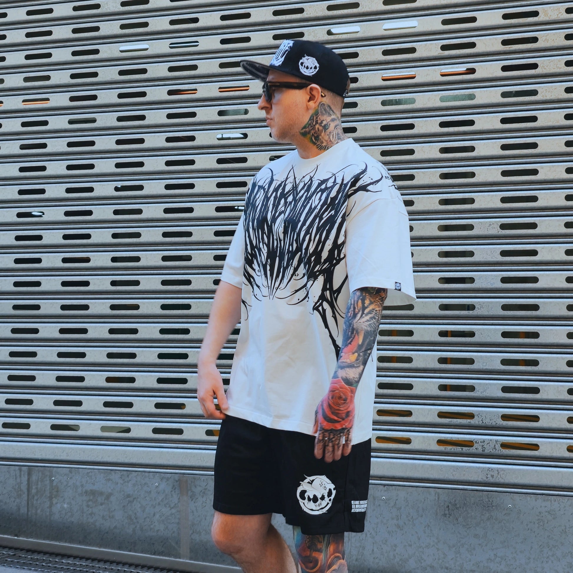 Man wearing a white t-shirt with black graphics and black shorts, standing in front of a metallic shutter.