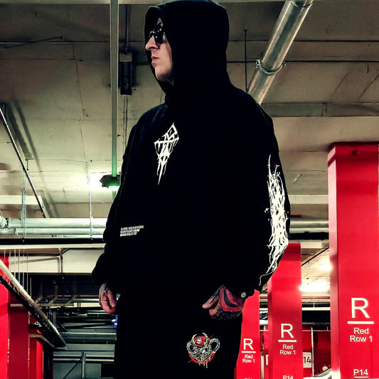 A person wearing a black hooded sweatshirt and matching black pants stands in a parking structure. The hoodie has white graphic prints on the chest, sleeve, and lower front, and the pants feature a small red and gold crest graphic near the lower left leg.