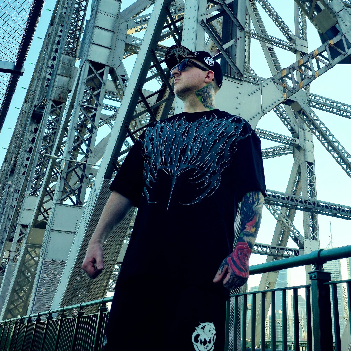 Man wearing a black t-shirt with a unique design in front of a large metal bridge.
