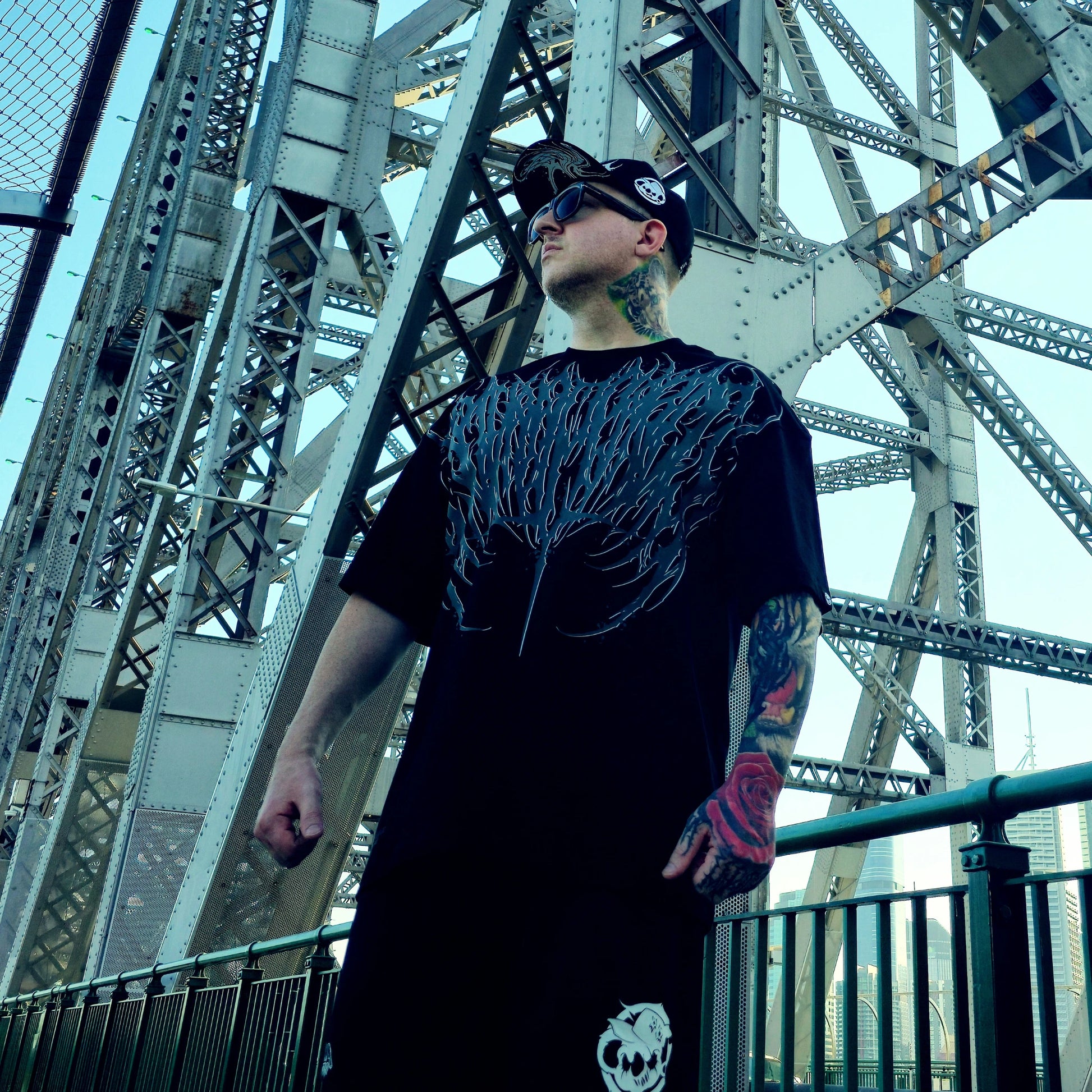 Man wearing a black t-shirt with a unique design in front of a large metal bridge.