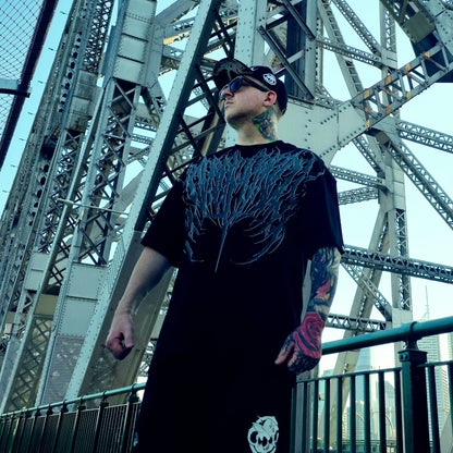 Man wearing a black t-shirt with a unique design in front of a large metal bridge.