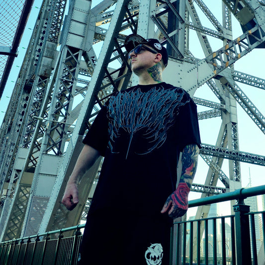 Man wearing a black t-shirt with a unique design in front of a large metal bridge.