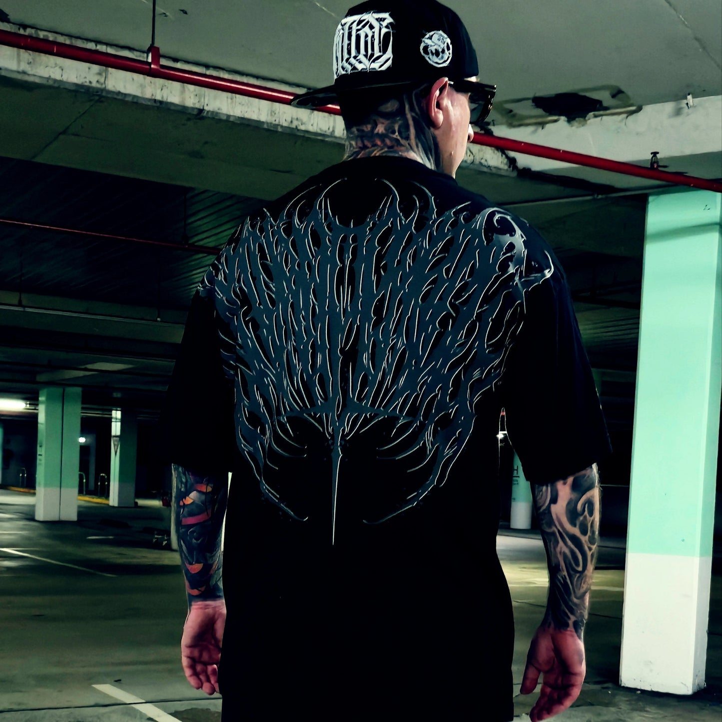 A tattooed man viewed from behind wearing a black t-shirt with a large, detailed black and gray wing-like graphic printed across the entire back, paired with a black baseball cap in a concrete parking garage.