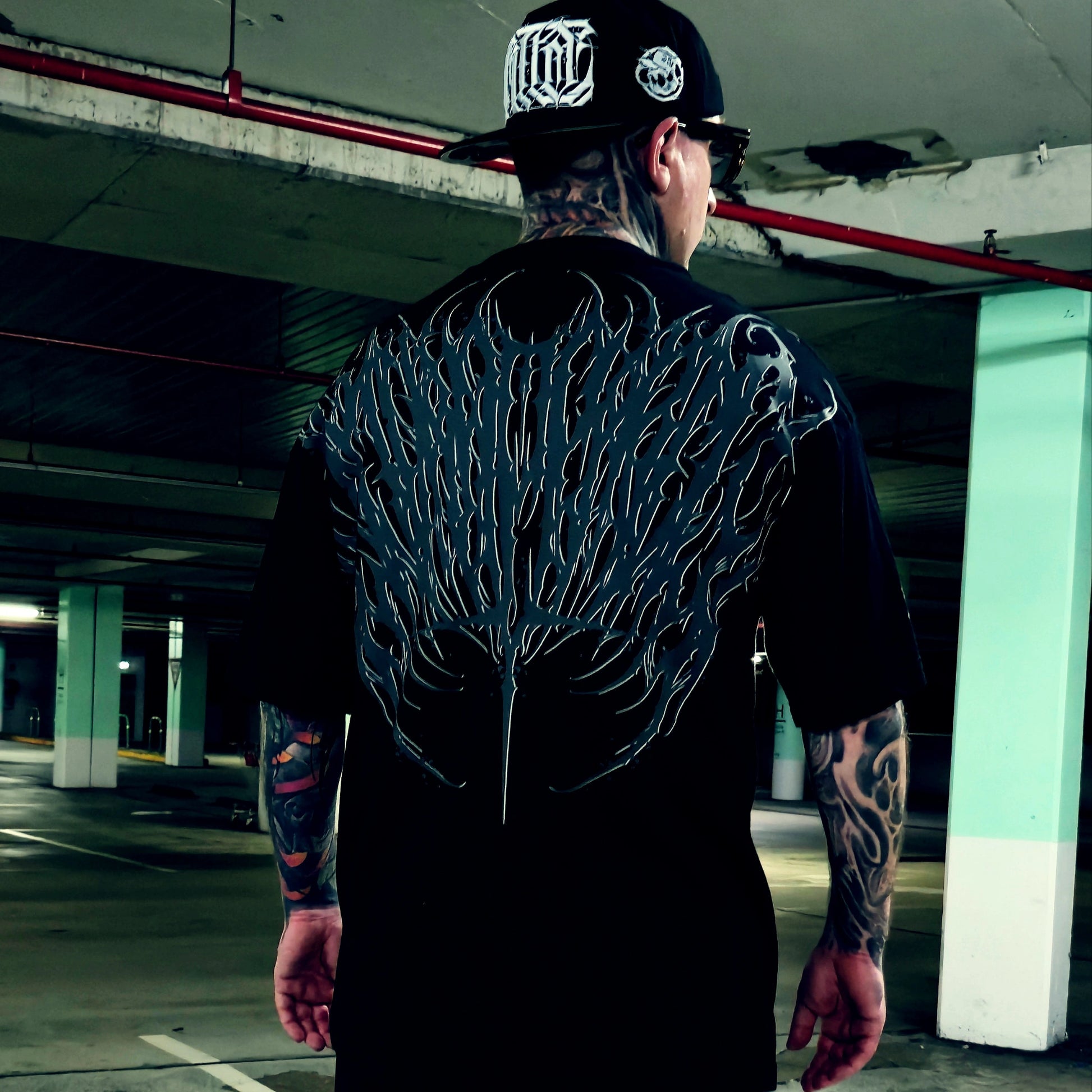 A tattooed man viewed from behind wearing a black t-shirt with a large, detailed black and gray wing-like graphic printed across the entire back, paired with a black baseball cap in a concrete parking garage.