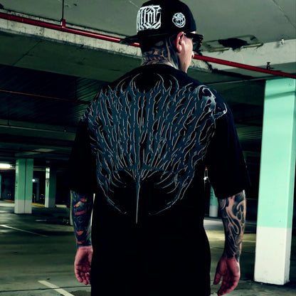 A tattooed man viewed from behind wearing a black t-shirt with a large, detailed black and gray wing-like graphic printed across the entire back, paired with a black baseball cap in a concrete parking garage.