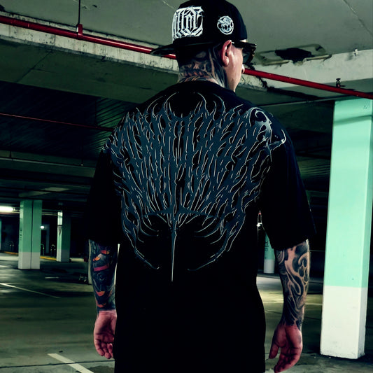 A tattooed man viewed from behind wearing a black t-shirt with a large, detailed black and gray wing-like graphic printed across the entire back, paired with a black baseball cap in a concrete parking garage.