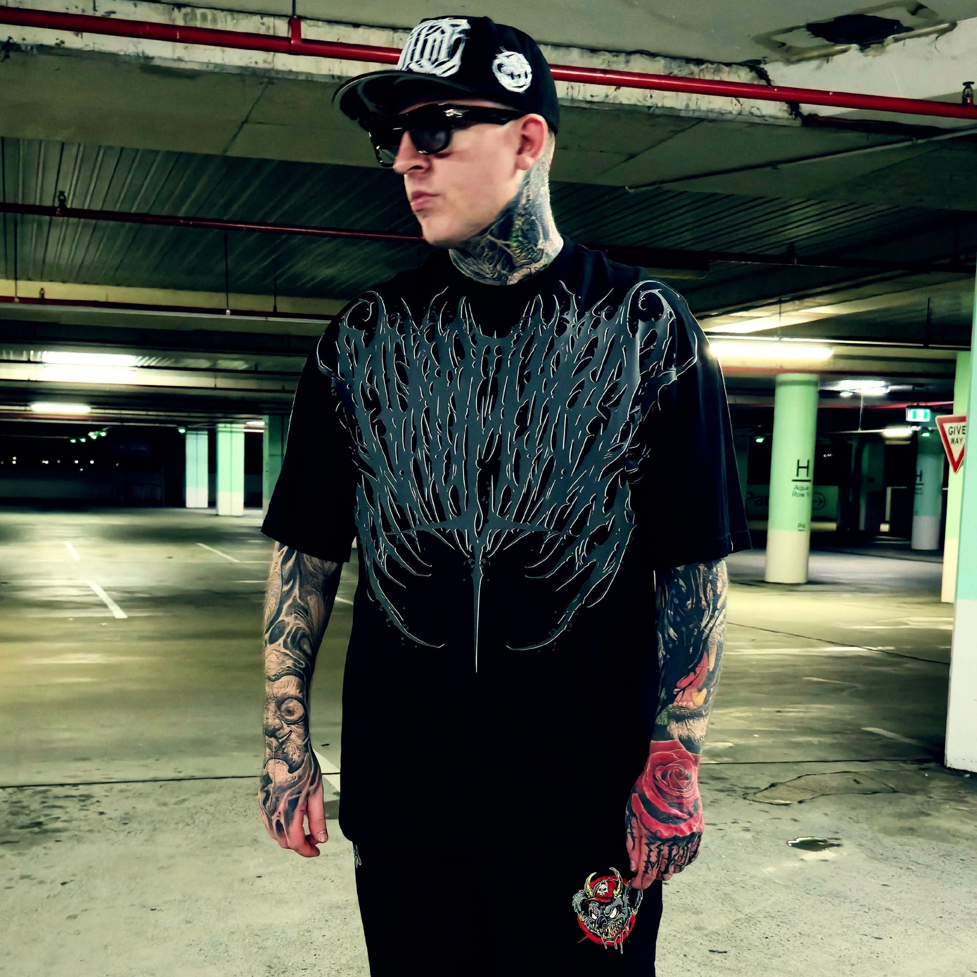 A tattooed man wearing a black band t-shirt with a large gray tree graphic, a black baseball cap, and sunglasses, standing in a concrete parking garage.