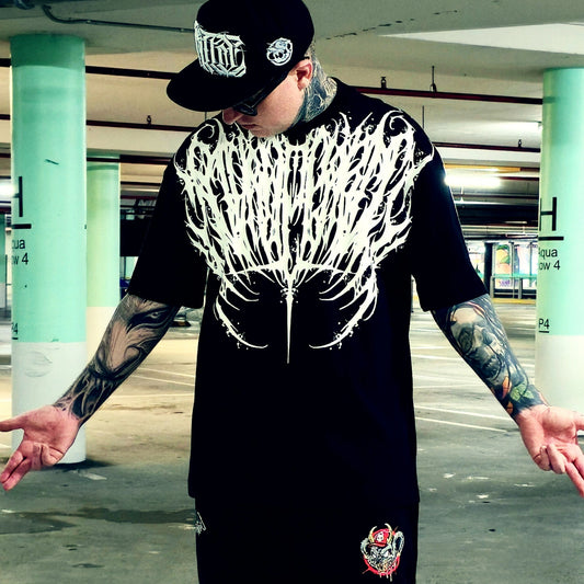 A tattooed man wearing a black t-shirt with a large white wing graphic on the front, a black baseball cap, and sunglasses, standing in a parking garage with his arms outstretched.