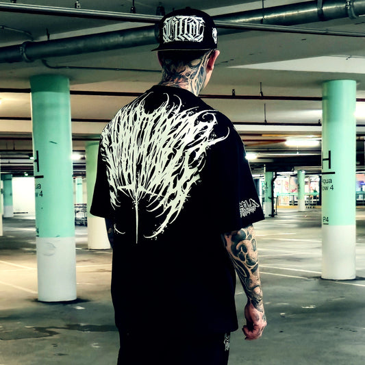 A tattooed man viewed from behind in a parking garage, wearing a black t-shirt with a large white wing-like graphic on the back and a black baseball cap with white lettering.