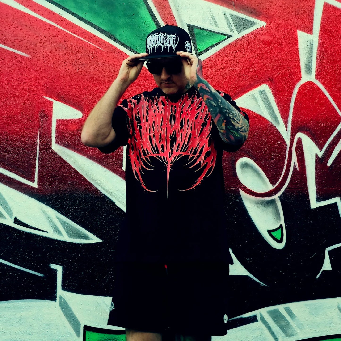 Person wearing a black t-shirt with red graphic design and cap, standing in front of a colorful graffiti wall.