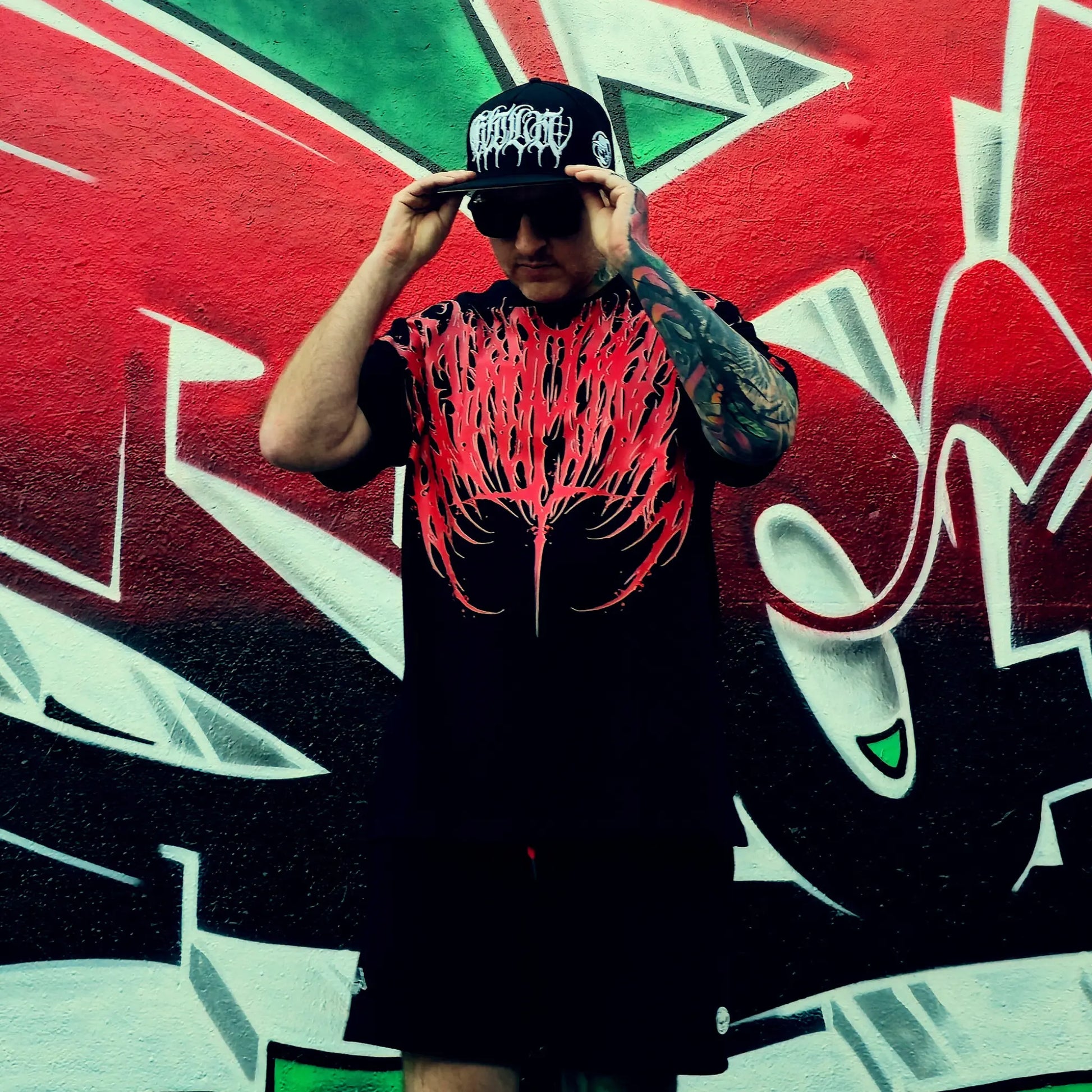 Person wearing a black t-shirt with red graphic design and cap, standing in front of a colorful graffiti wall.