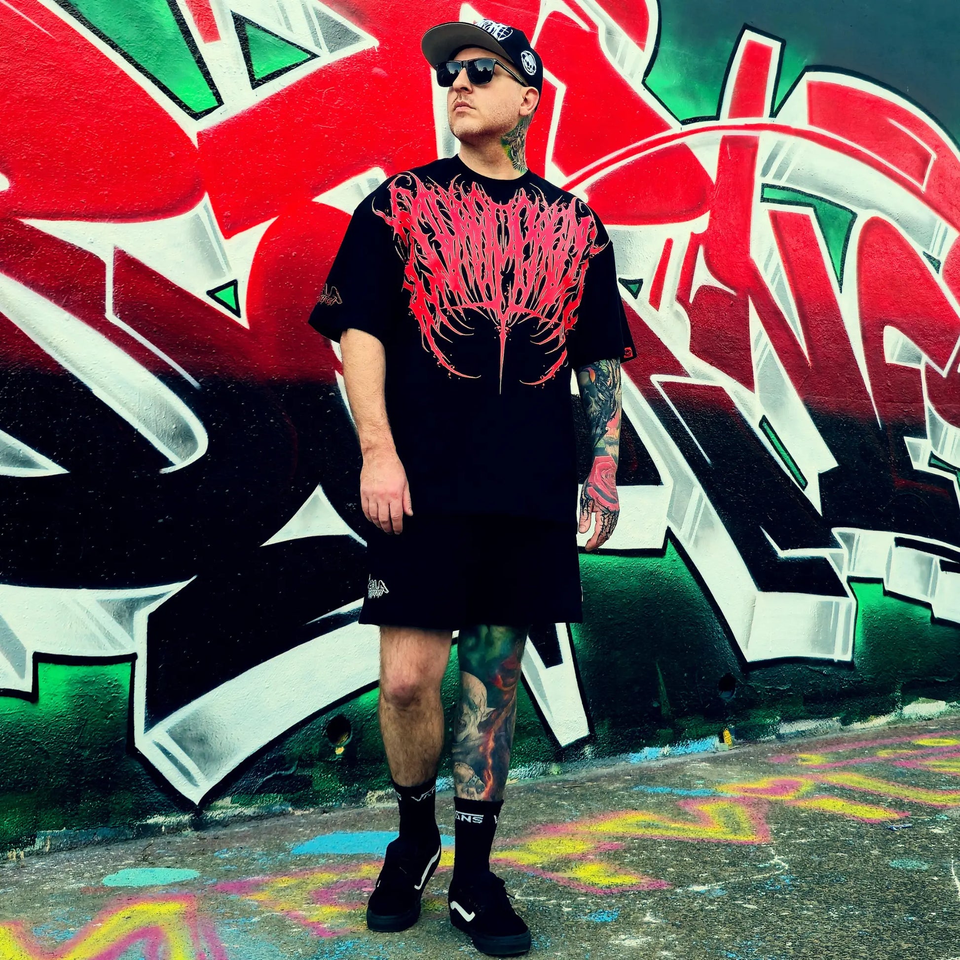 Person wearing a black t-shirt with red design and black shorts standing in front of colorful graffiti.