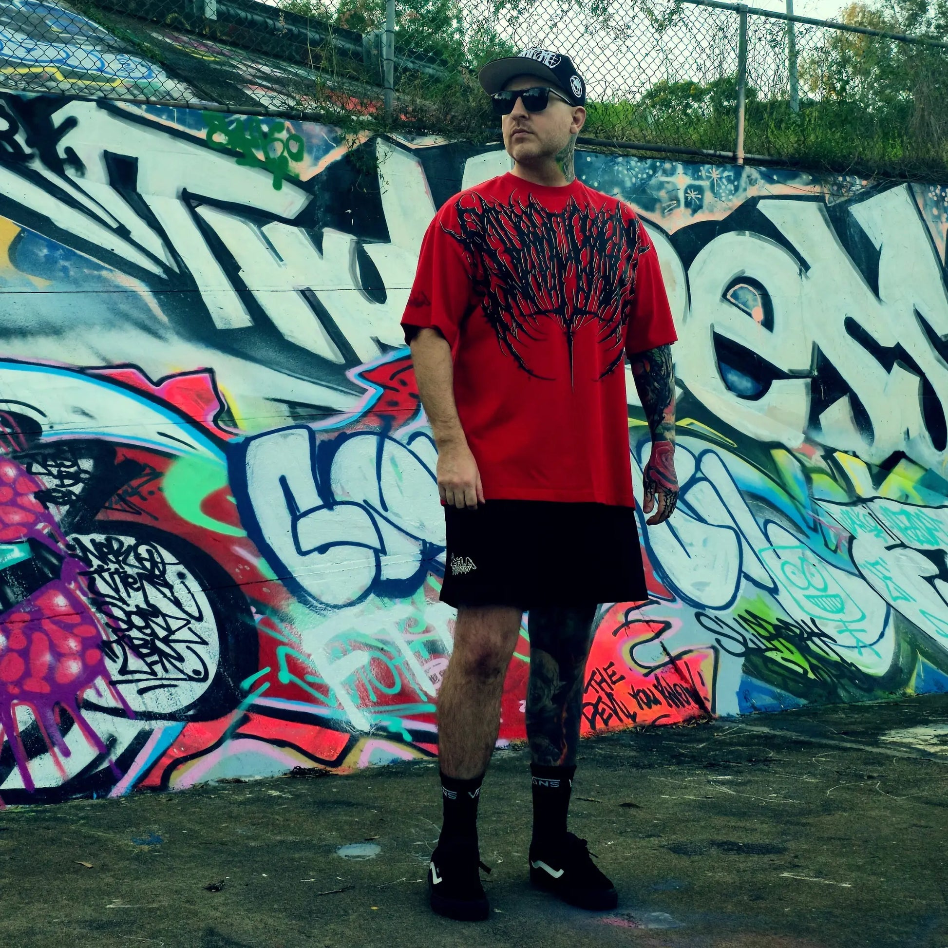 Man wearing a red shirt with black graphics and black shorts standing in front of a colorful graffiti wall.