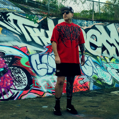 Man wearing a red shirt with black graphics and black shorts standing in front of a colorful graffiti wall.