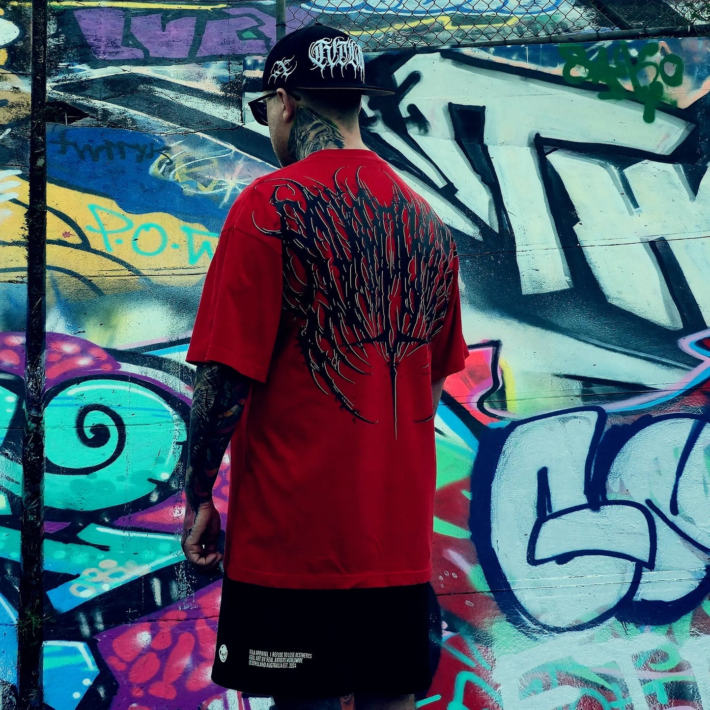 Person wearing a red t-shirt with a black design on the back, standing in front of a colorful graffiti wall.