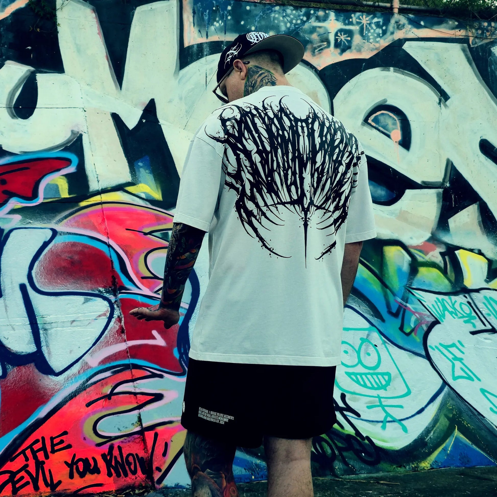 Person wearing a white t-shirt with black graphics in front of a colorful graffiti wall.