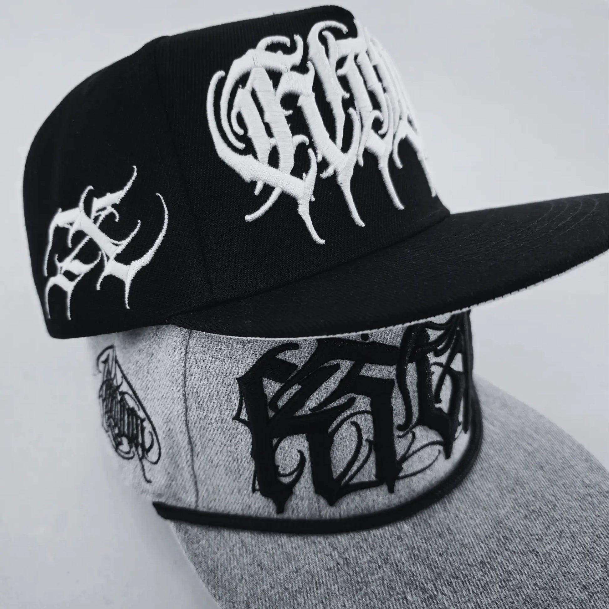 Two baseball caps with text patterns, one in black and the other in grey, both with stylized text designs. RTLA-Apparel