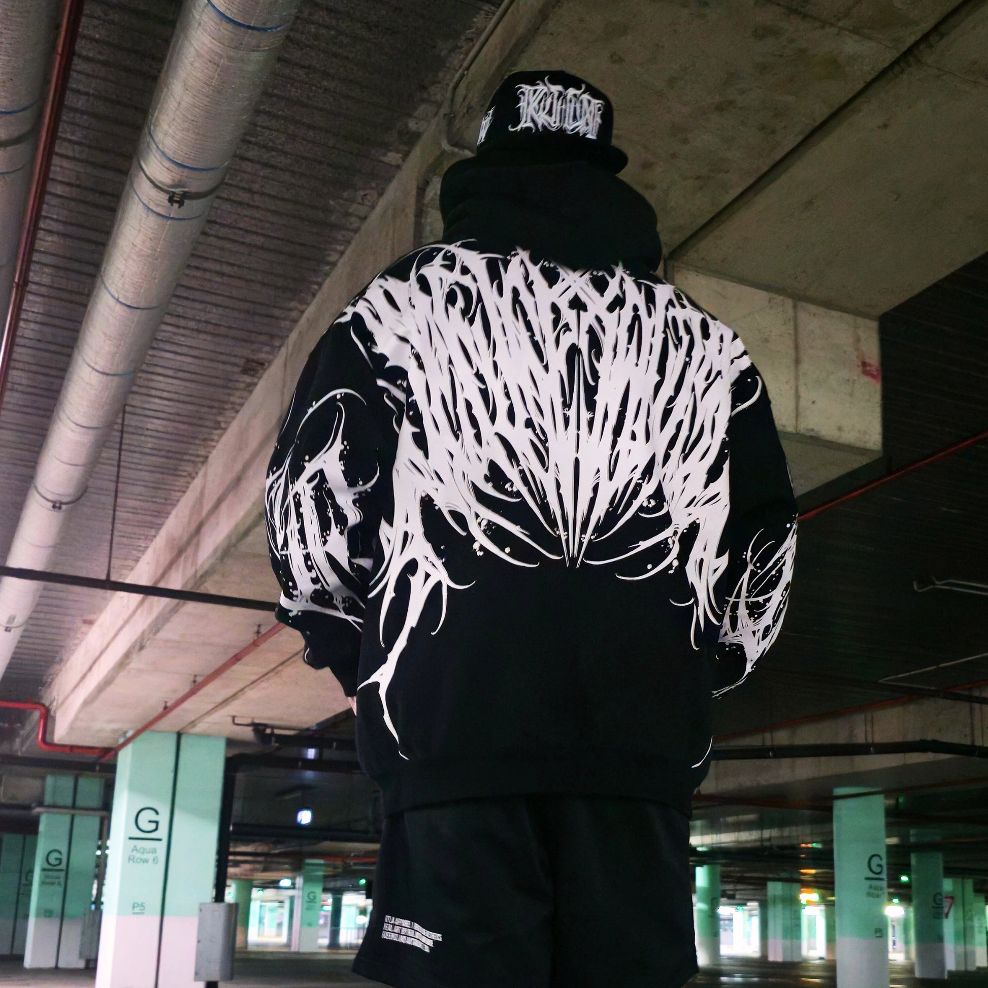 Person wearing a black hoodie with white abstract design in an underground parking garage.