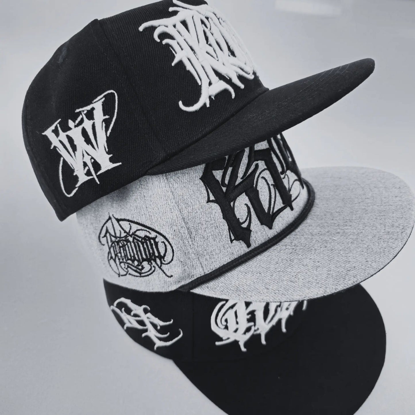 Two snapback caps with embroidered text designs, one primarily black and the other primarily grey, both with 'TRIPLE CROWN' written on them. RTLA-Apparel