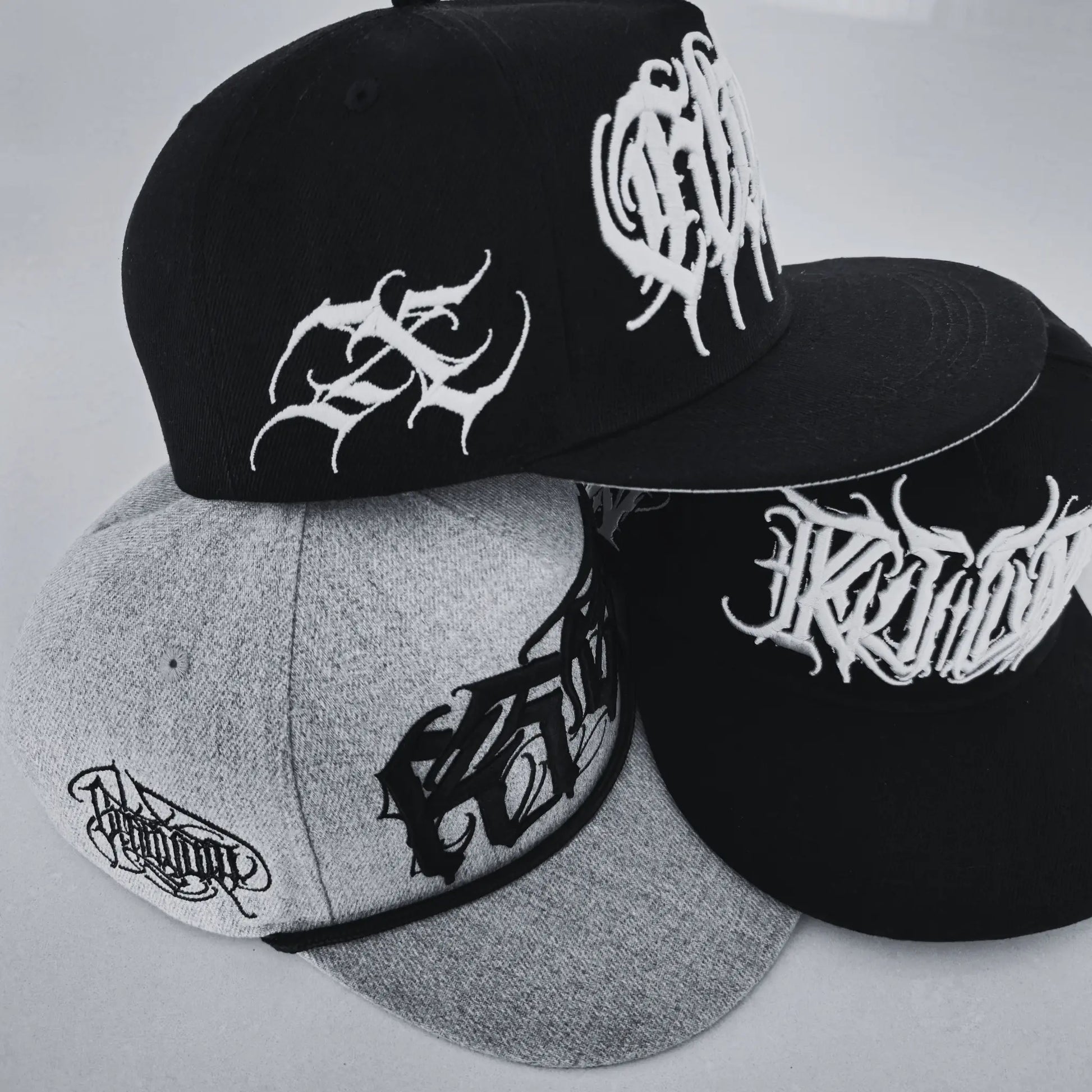 Three caps in black and grey with white logo on a light background. RTLA-Apparel