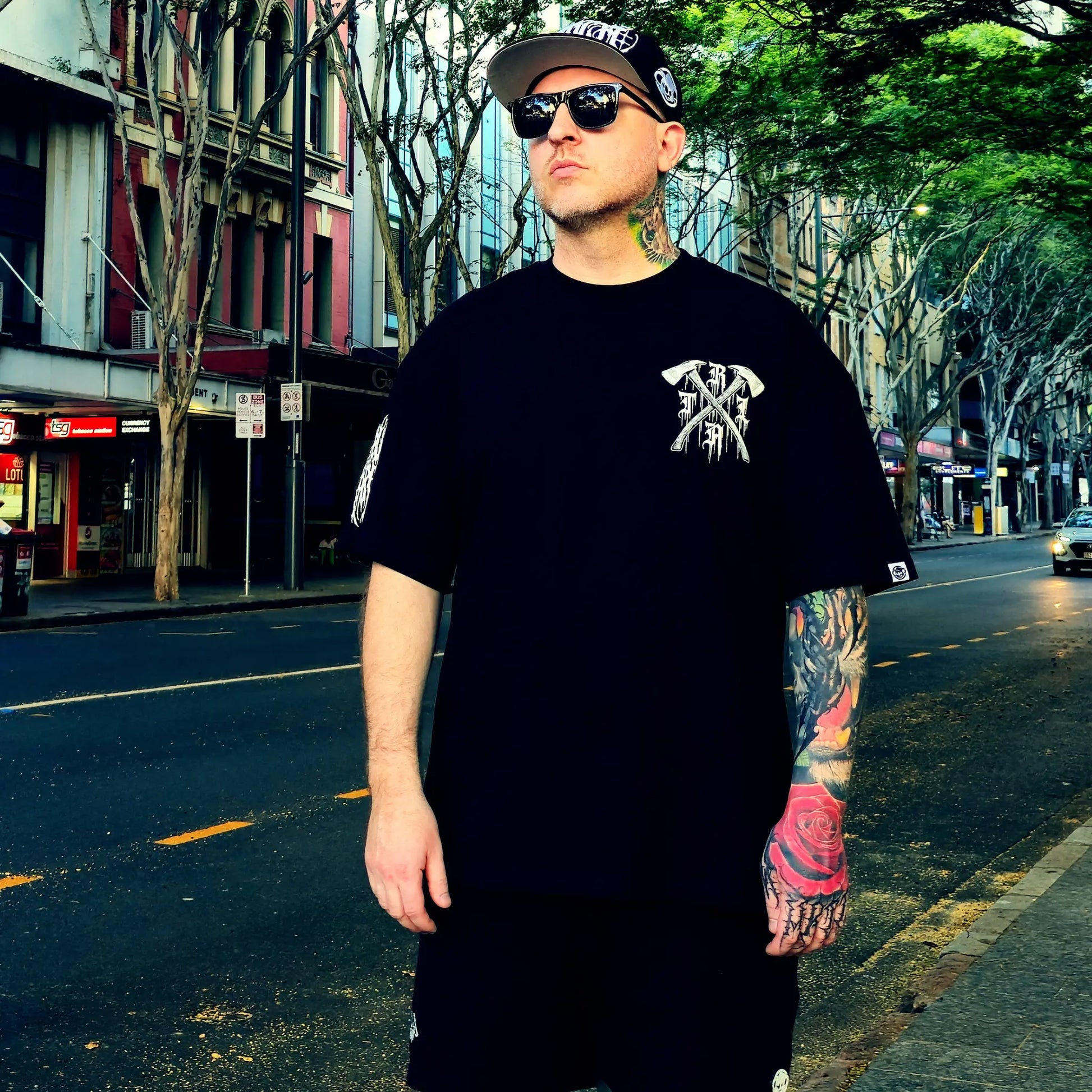 Man wearing a black t-shirt with a logo and sunglasses on a city street.