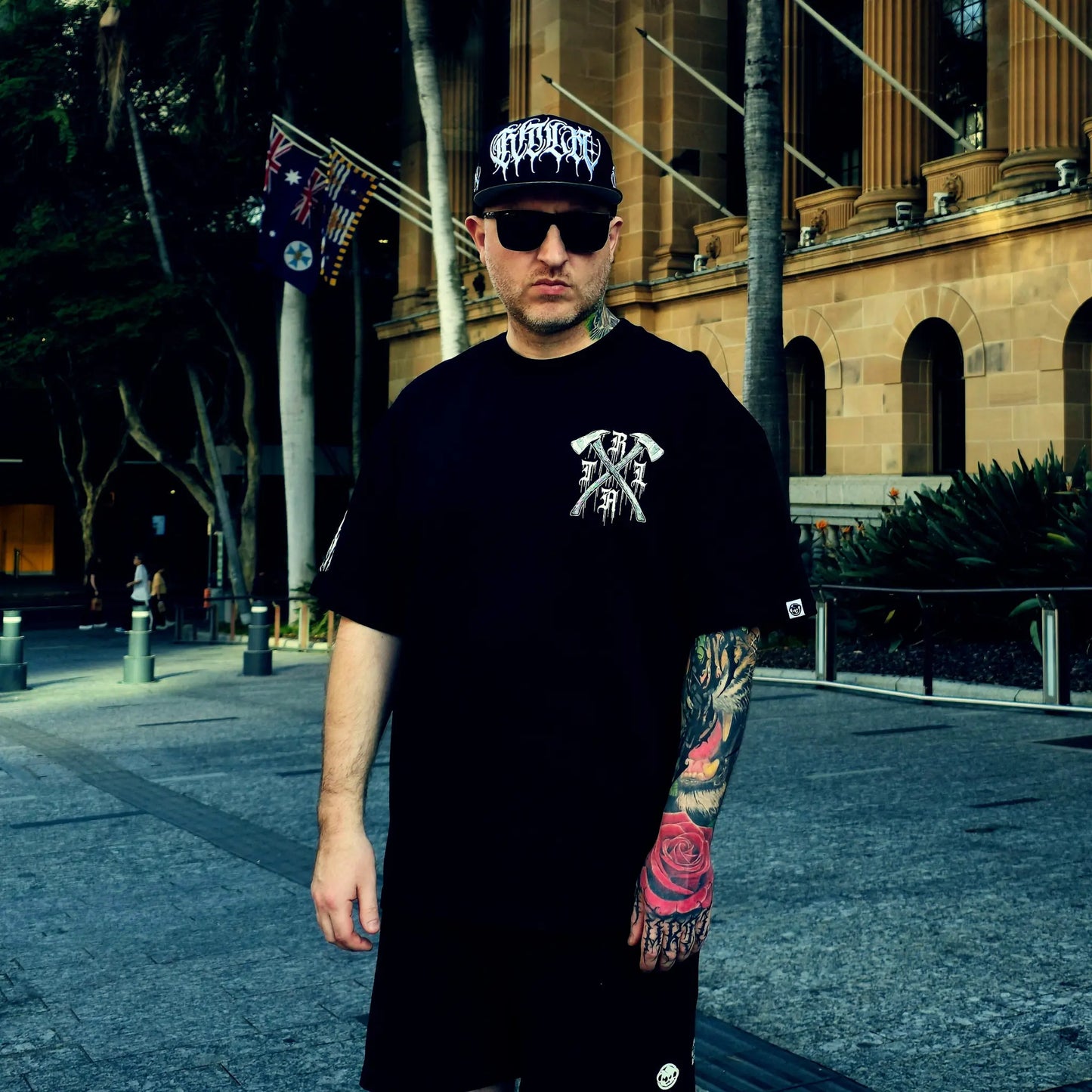 Man wearing a black t-shirt with a logo and a cap, standing in an urban setting.