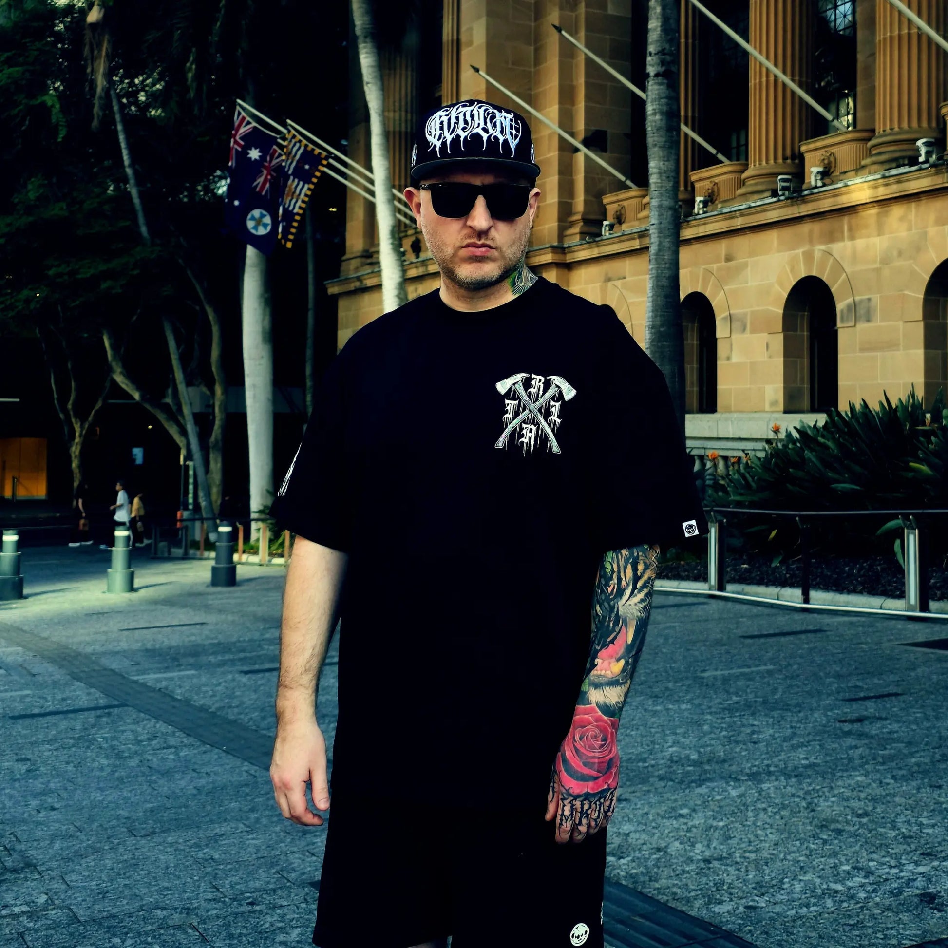 Man wearing a black t-shirt with a logo and a cap, standing in an urban setting.