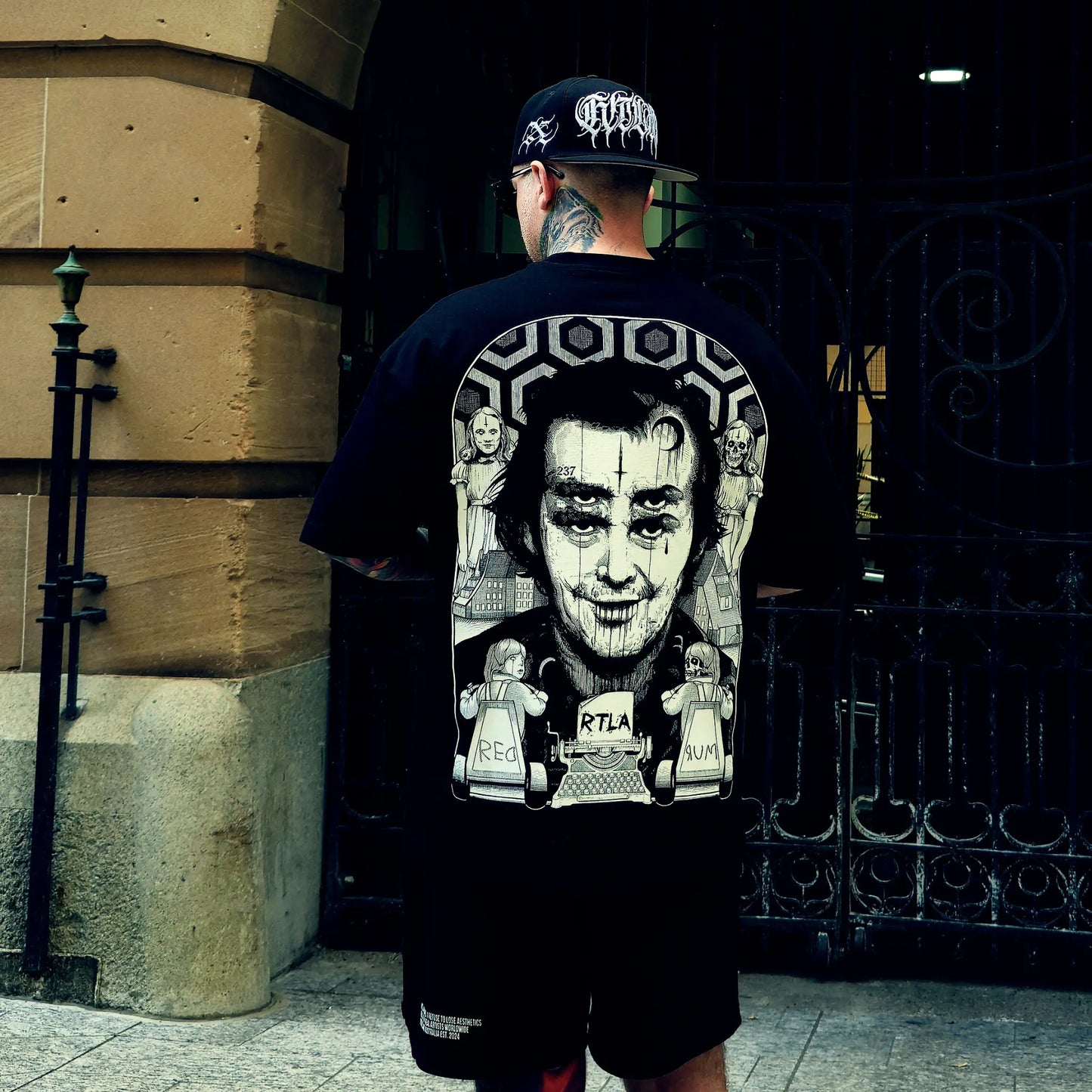 Person wearing a black t-shirt with graphic design and a cap, standing in front of a building entrance.