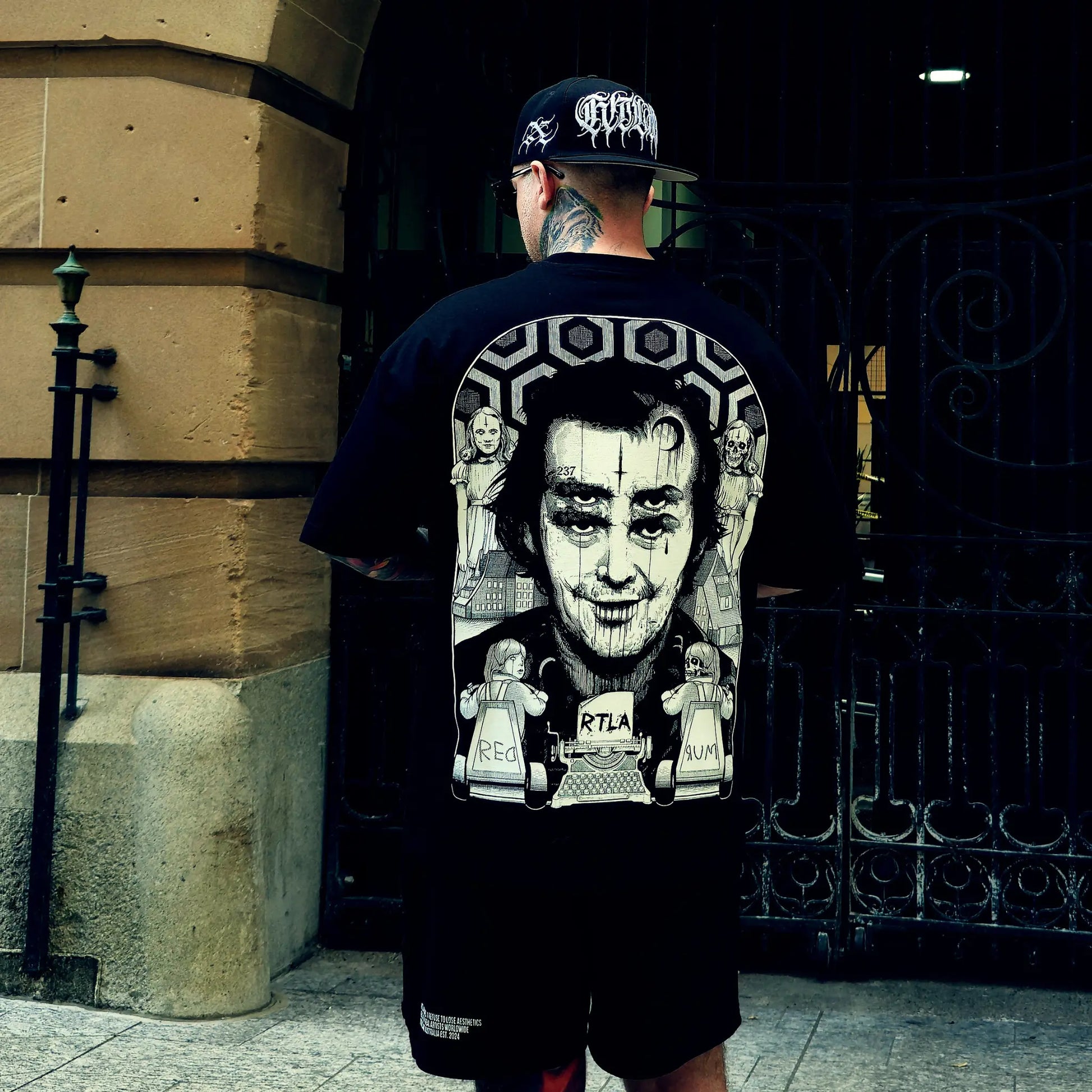 Person wearing a black t-shirt with graphic design and a cap, standing in front of a building entrance.