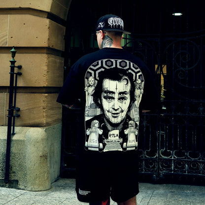 Person wearing a black t-shirt with graphic design and a cap, standing in front of a building entrance.