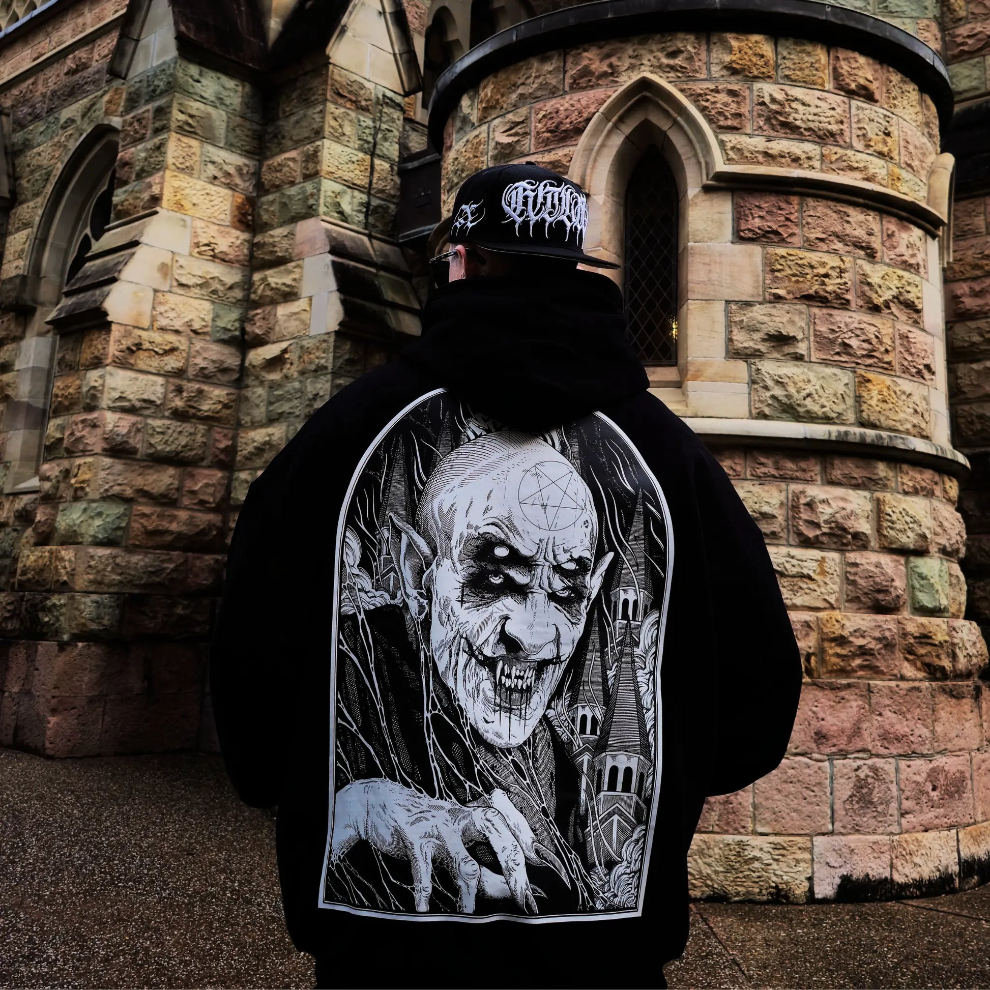 Person wearing a black hoodie with a graphic design of a skeleton holding a skull, standing in front of a stone building. RTLA-Apparel