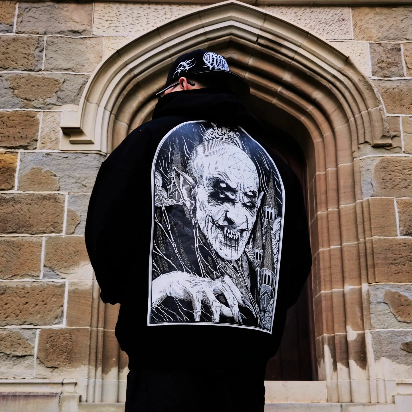A person wearing a black hoodie with a white graphic of a character on the back, standing in front of a stone archway. RTLA-Apparel