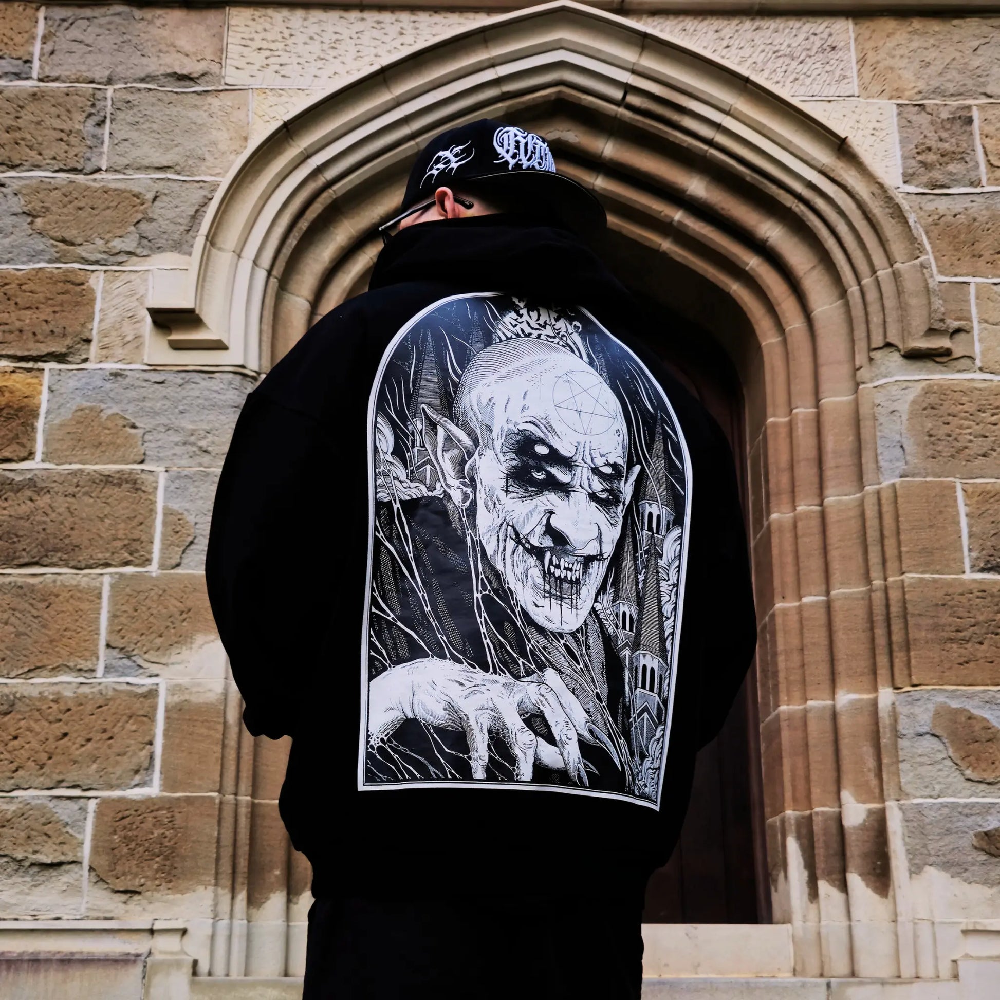 A person wearing a black hoodie with a white graphic of a character on the back, standing in front of a stone archway. RTLA-Apparel