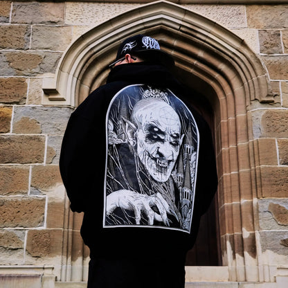 A person wearing a black hoodie with a white graphic of a character on the back, standing in front of a stone archway. RTLA-Apparel