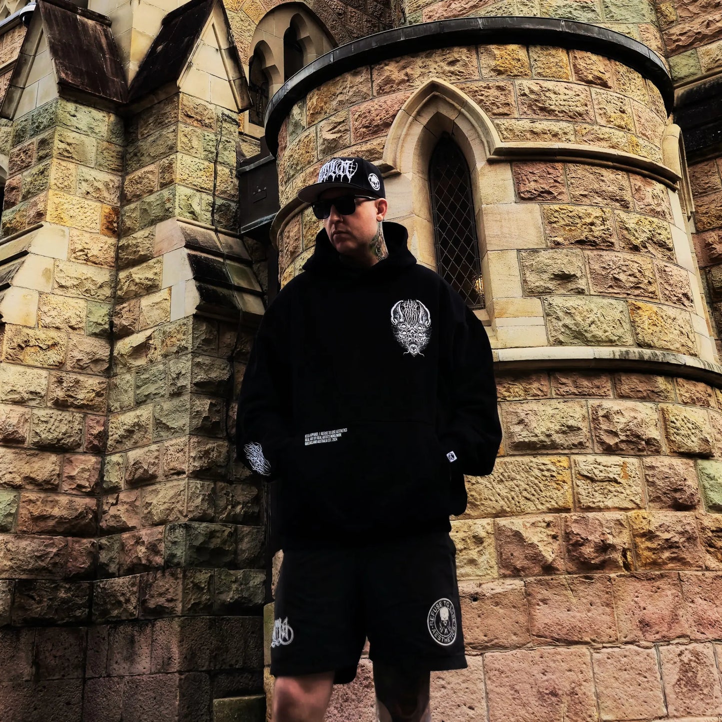 Person wearing a black hoodie and shorts with logos, standing in front of a stone building. RTLA-Apparel