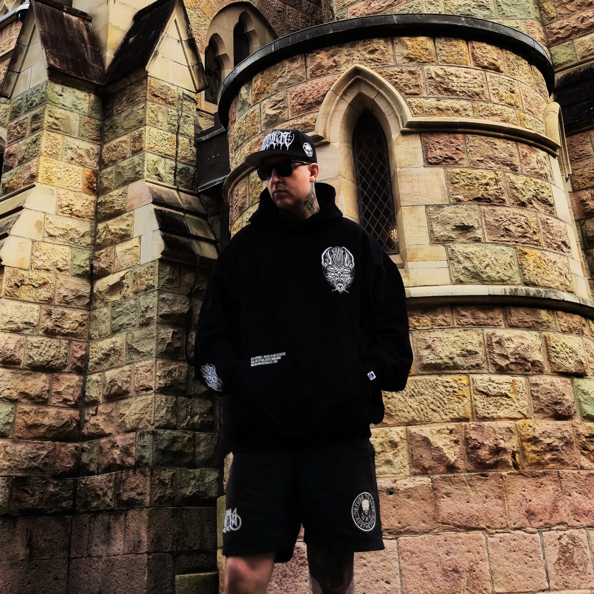 Person wearing a black hoodie and shorts with logos, standing in front of a stone building. RTLA-Apparel
