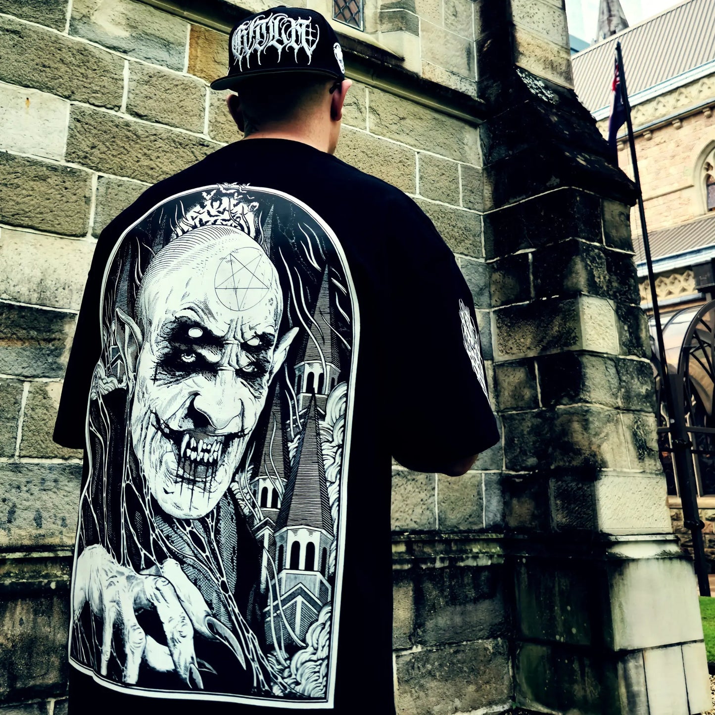 Person wearing a black t-shirt with a graphic design of a skeleton and castle, standing in front of a stone building. RTLA-Apparel