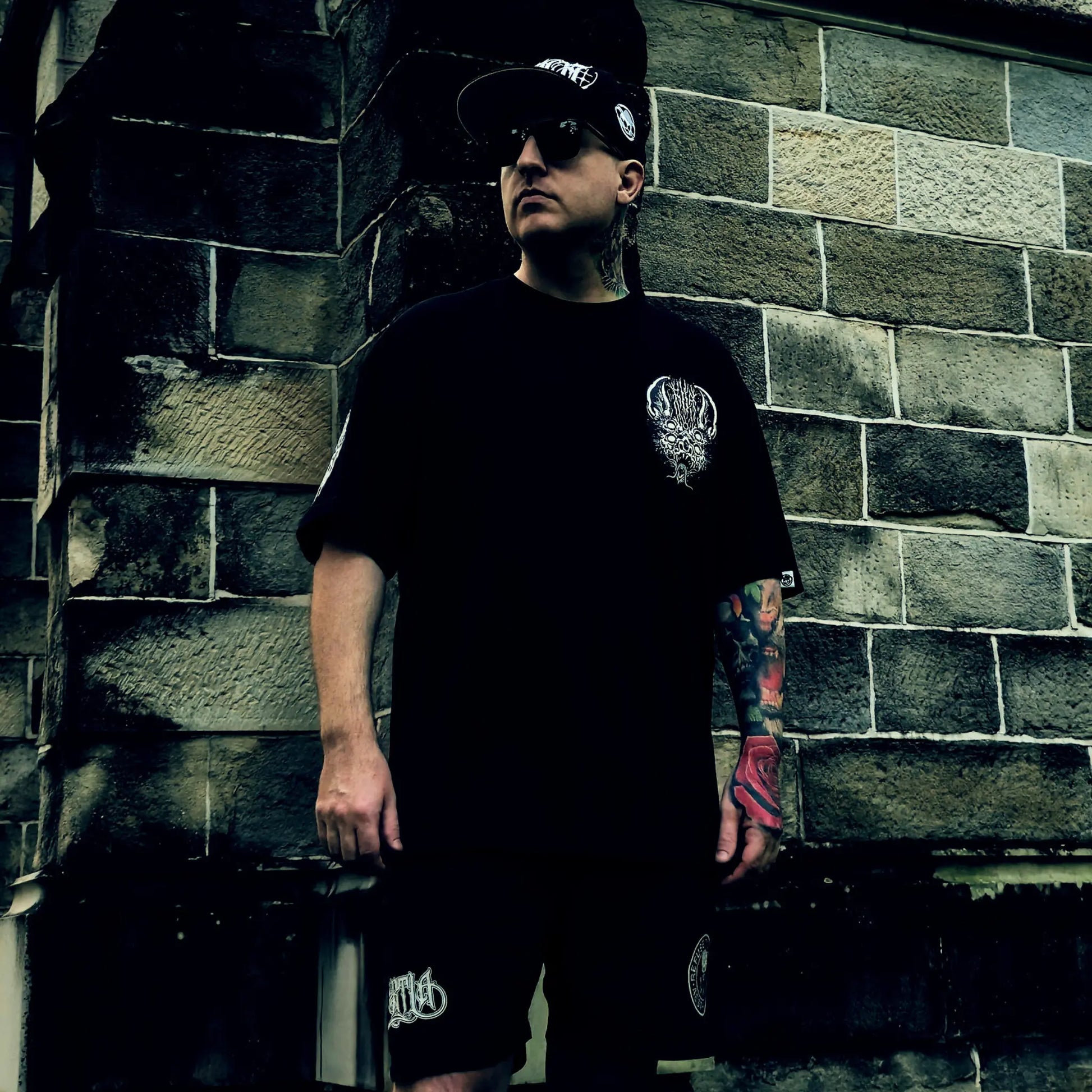 Man wearing a black t-shirt and shorts with a tattooed arm, standing against a brick wall. RTLA Apparel