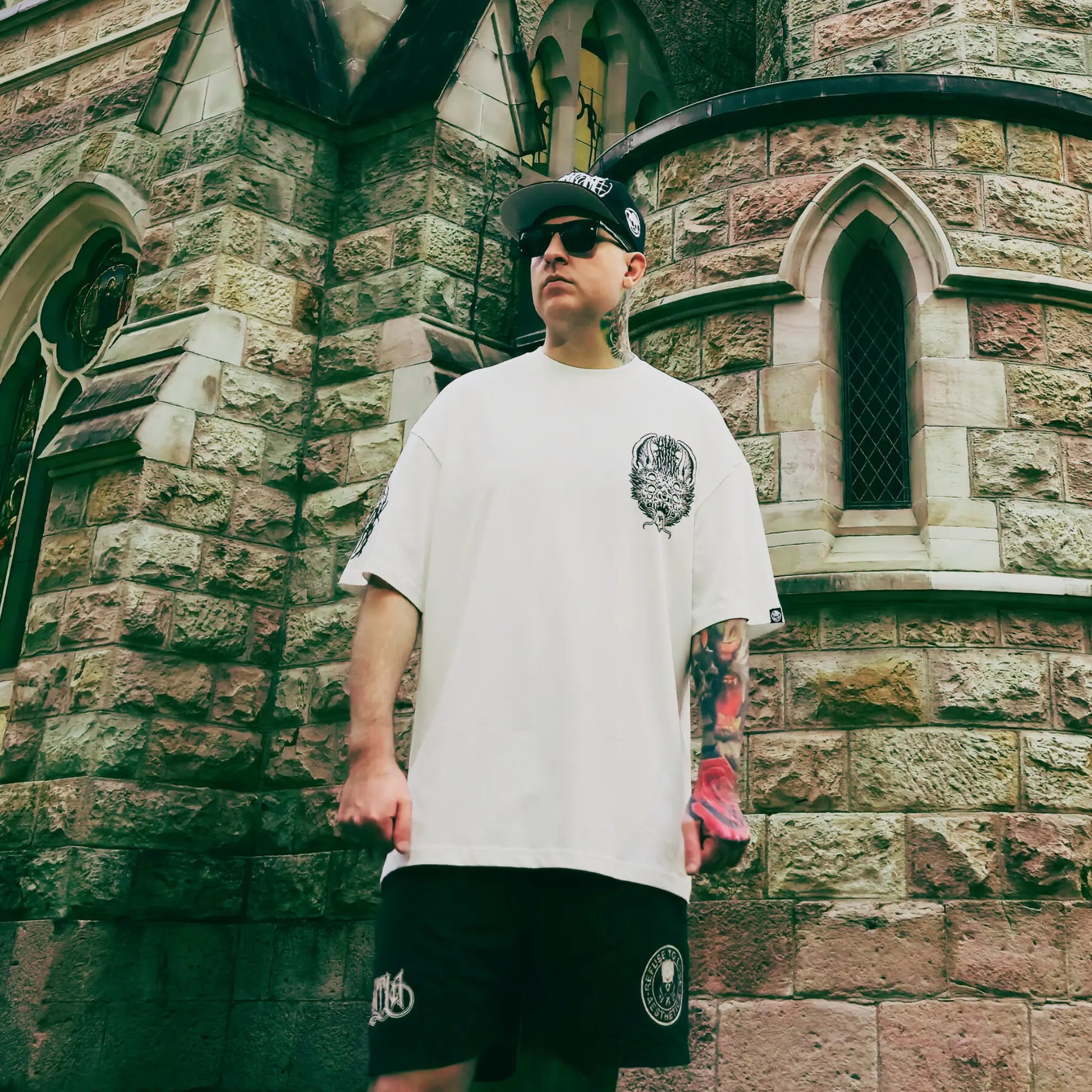 Man wearing a white t-shirt and black shorts with a logo, standing in front of a stone building. RTLA Apparel