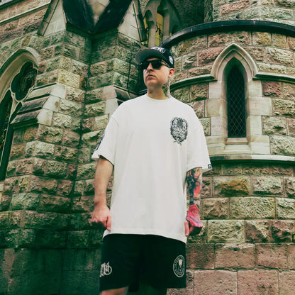 Man wearing a white t-shirt and black shorts with a logo, standing in front of a stone building. RTLA Apparel
