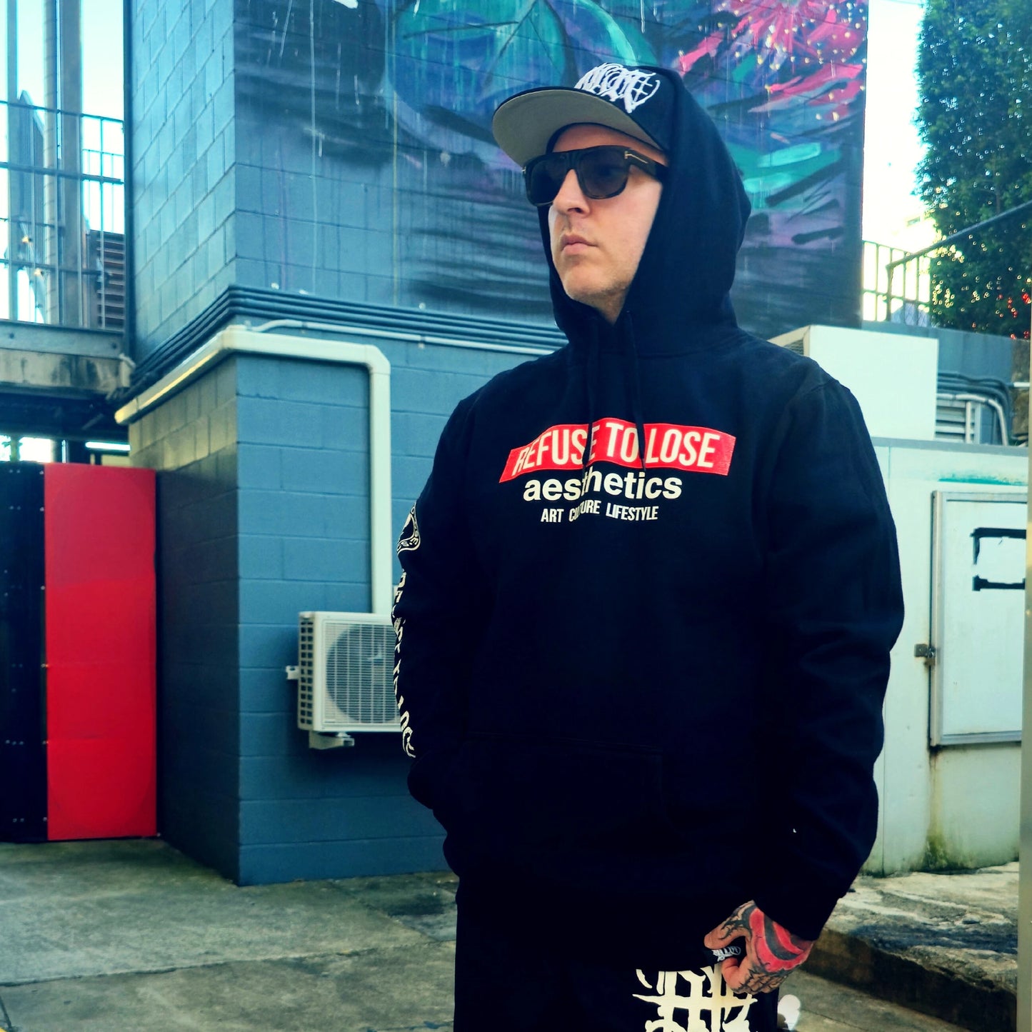 Person wearing a black hoodie with text, sunglasses, and a cap in an urban setting.