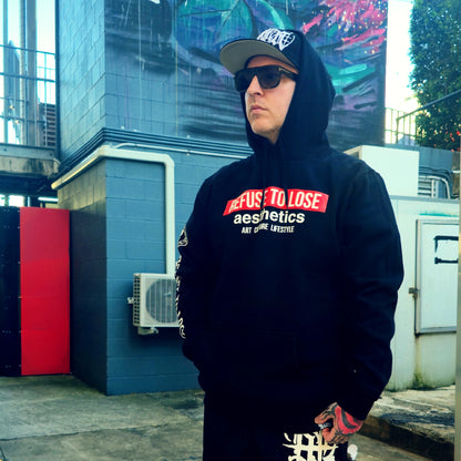 Person wearing a black hoodie with text, sunglasses, and a cap in an urban setting.
